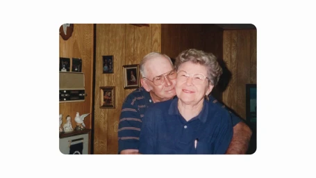 An elderly couple embracing indoors with wood paneled walls, small framed photos, and a figurine shelf in the background.