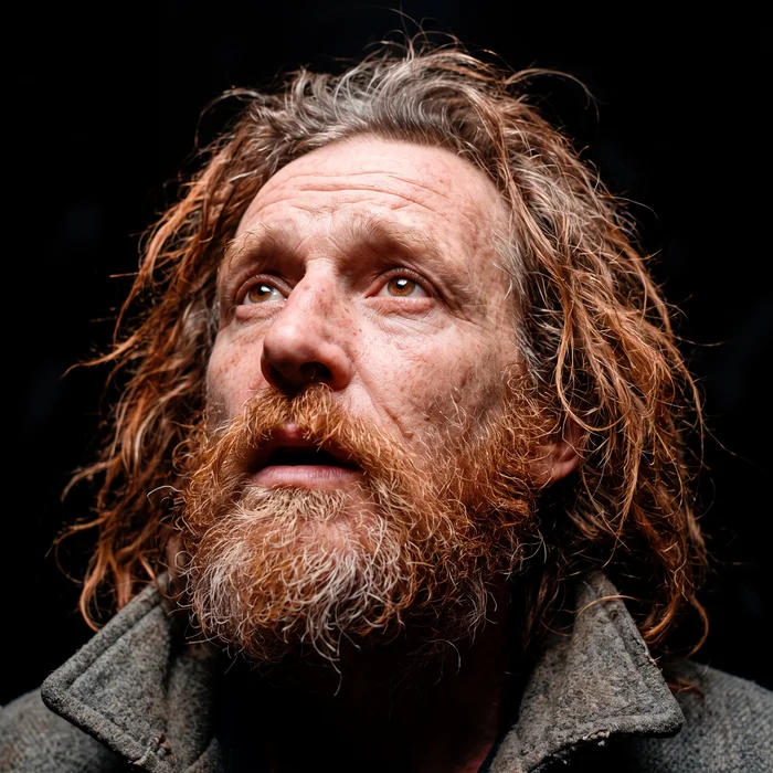 Close-up portrait of a man with long reddish-brown hair and a full beard looking upwards against a dark background