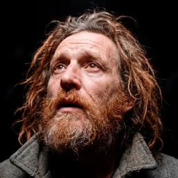 Close-up portrait of a man with long reddish-brown hair and a full beard looking upwards against a dark background