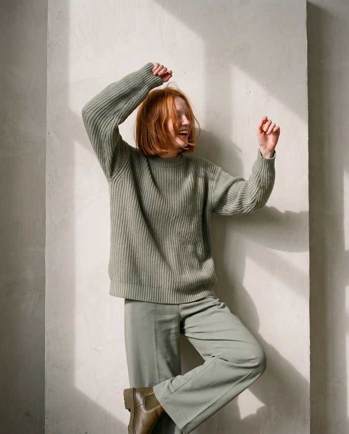 Red-haired woman wearing sage green knit sweater and matching trousers posing against a concrete wall