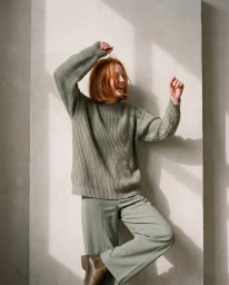 Red-haired woman wearing sage green knit sweater and matching trousers posing against a concrete wall