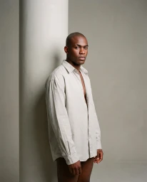 Black man with a shaved head wearing an oversized light grey buttoned shirt standing beside a large white pillar