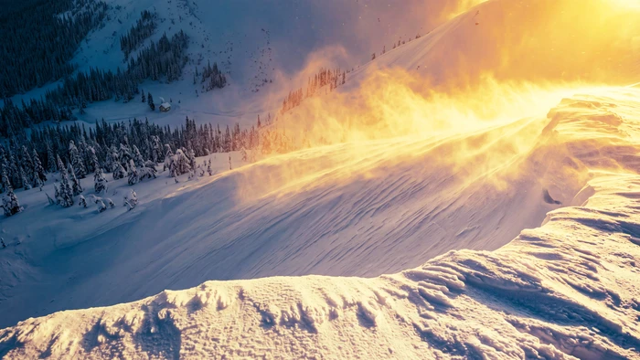 Aerial view of snow-covered mountain ridges and pine forests illuminated by golden sunrise light with blowing snow.