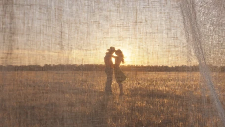 Silhouette of a man and woman embracing in a field at sunset, viewed through a sheer textured fabric curtain.