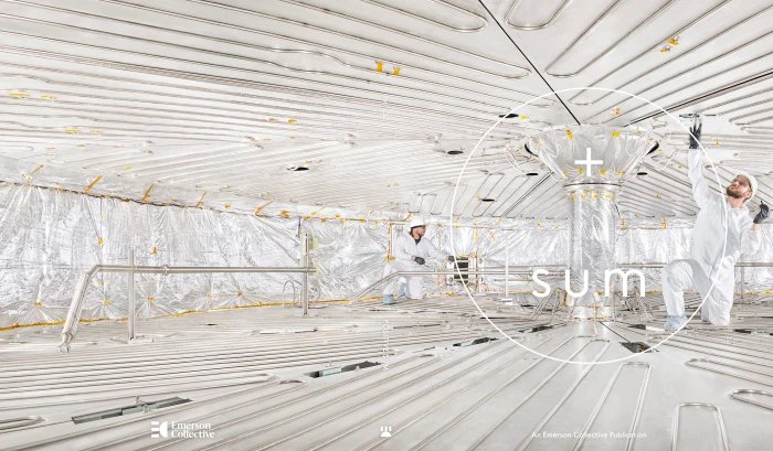 Two workers in white protective suits inside a large industrial facility with metallic reflective walls and floors.
