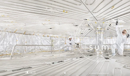 Two workers in white protective suits inside a large industrial facility with metallic reflective walls and floors.