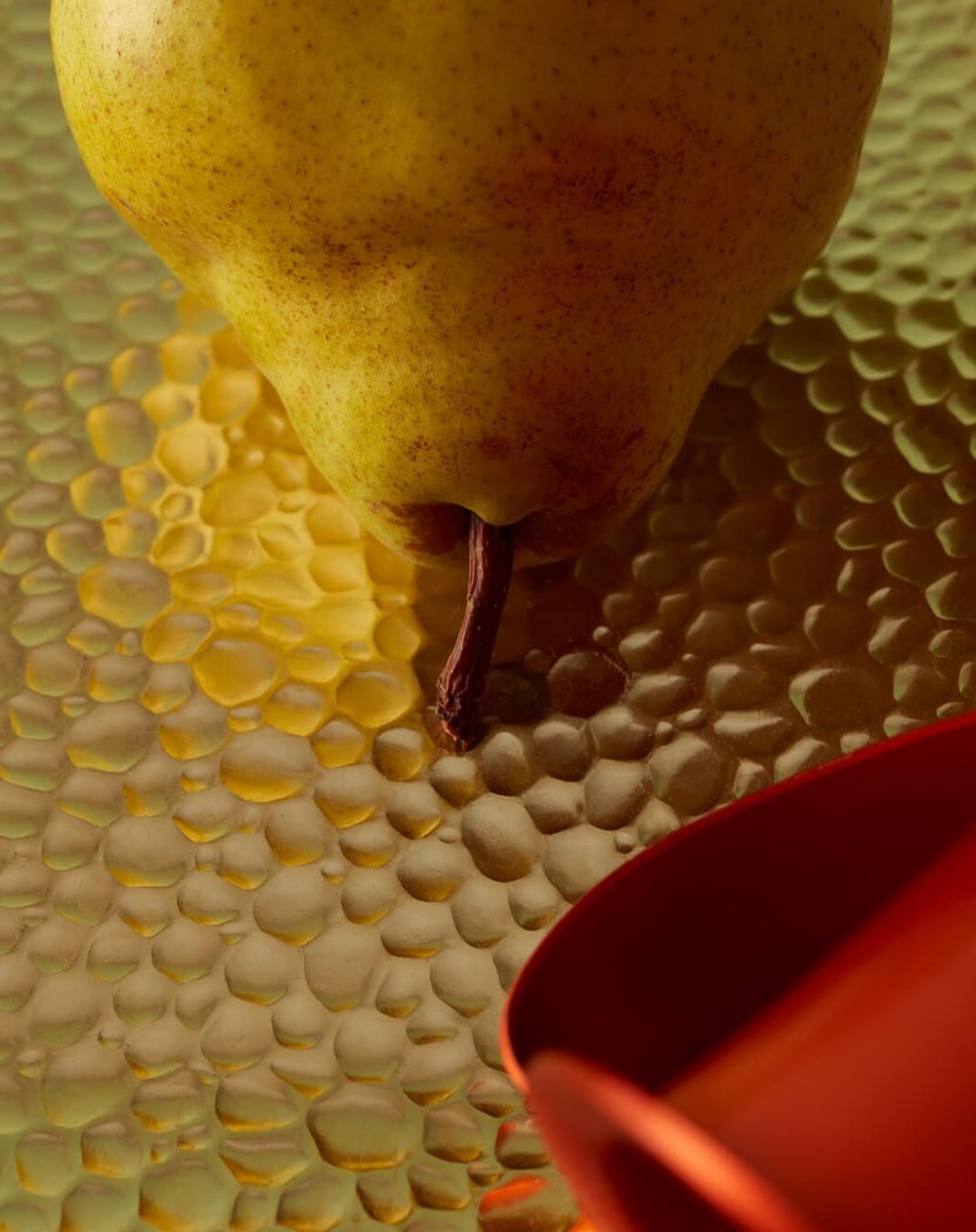 Close-up of a yellow pear with a red container on a textured surface, highlighting the fruit's shape and color contrasts.