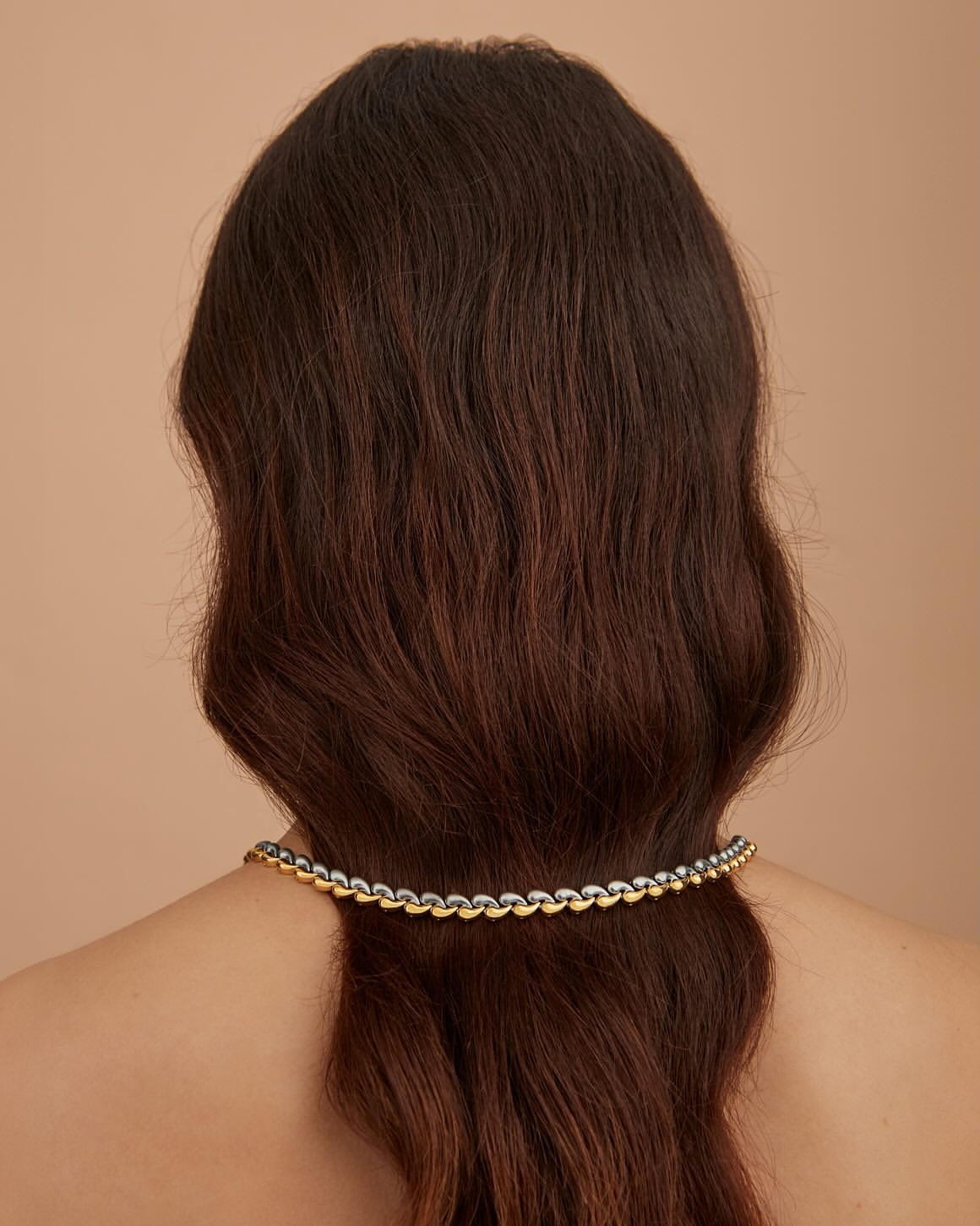 Brunette woman with long hair, viewed from behind, wearing a gold chain necklace against a soft beige background in a close-up shot.