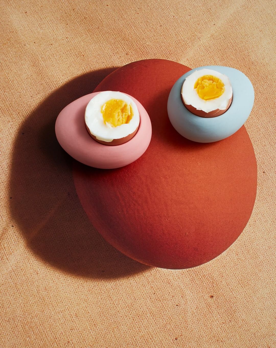 Still life featuring two pastel-colored egg cups holding halved boiled eggs atop a larger terracotta-colored egg on a fabric surface.