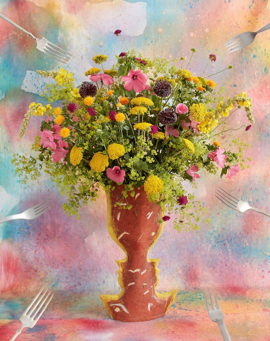 Vase with a lush flower arrangement featuring vibrant yellows, pinks, and greens, complemented by scattered forks and a watercolor backdrop.