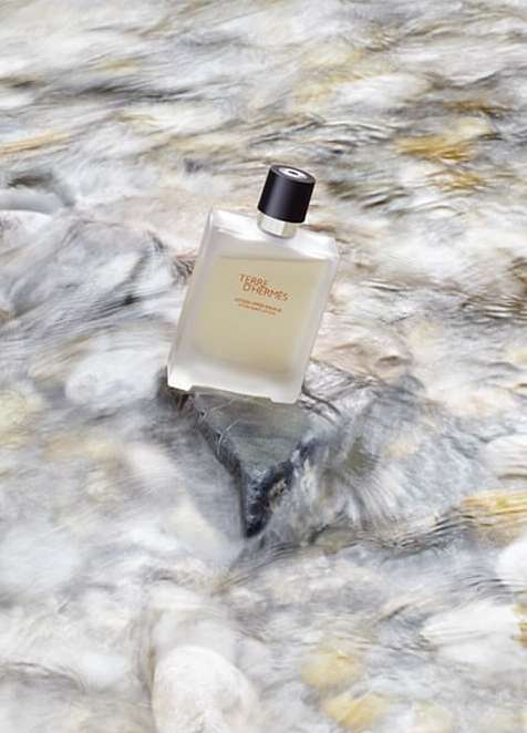 Frosted glass perfume bottle from Terre d'Hermes with a black cap is laying on water with rocks creating an alluring scene.
