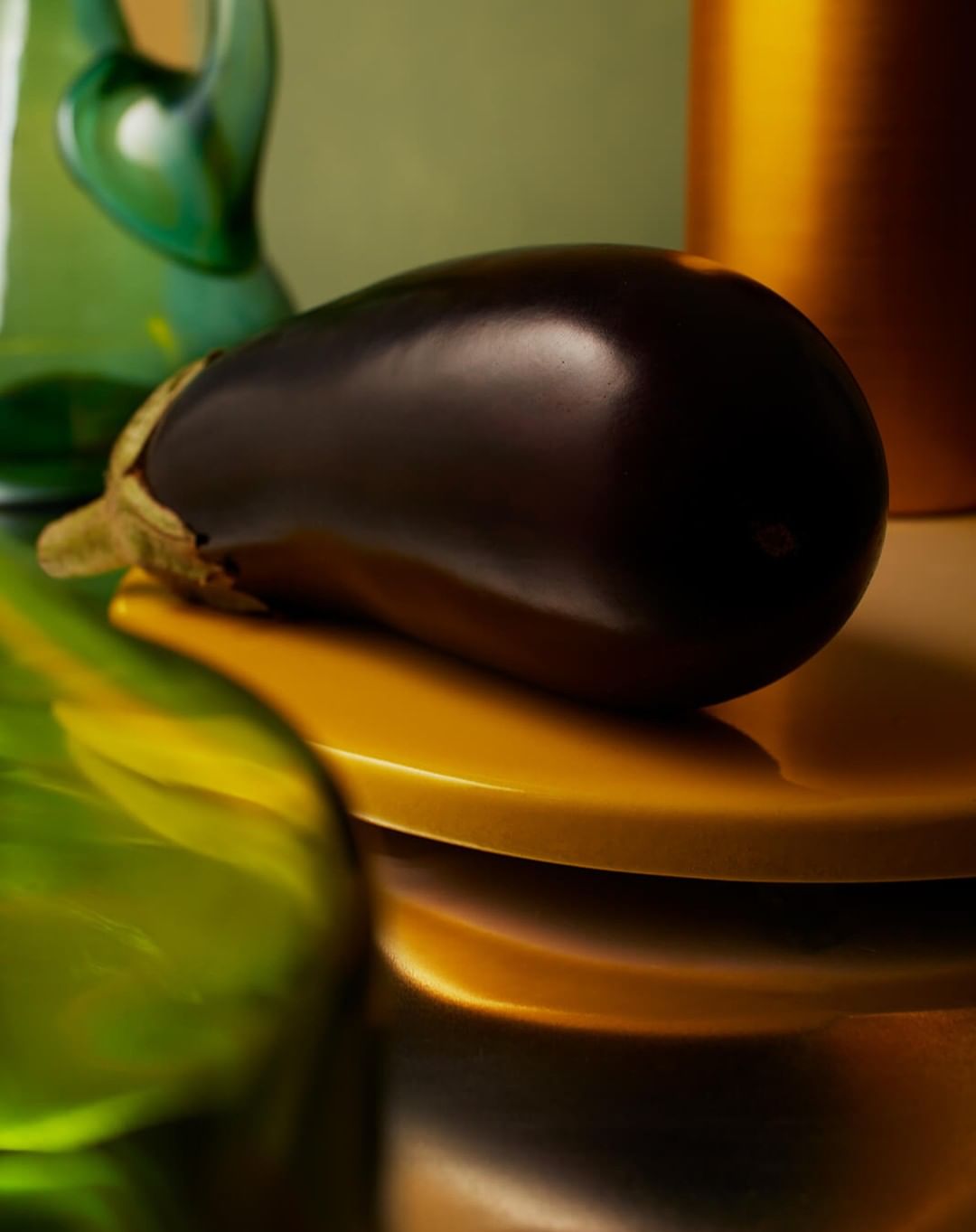 Still life shot of a glossy, dark purple eggplant on a yellow plate, with blurred green glass shapes and golden accents in the background.