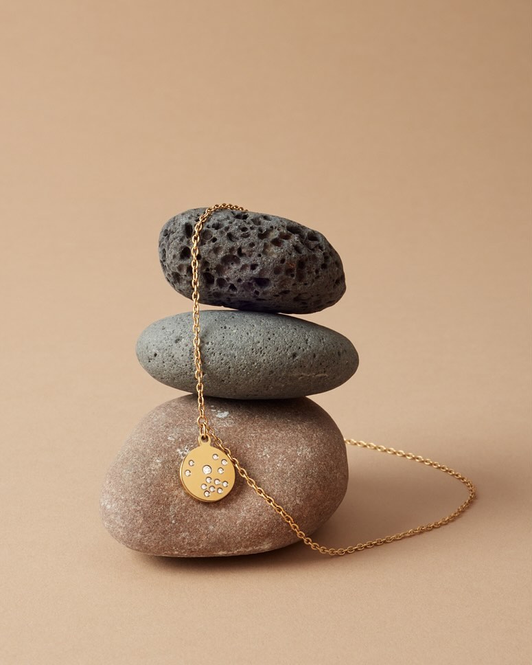Gold pendant necklace displayed on stacked stones against a beige background, minimalist jewellery still life, product shot.