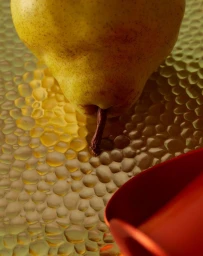 Close-up still life photograph of a fresh yellow pear resting on a textured golden glass surface with a red object in the foreground corner.