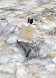 A translucent glass perfume bottle resting on a wet dark stone surrounded by rushing river water.