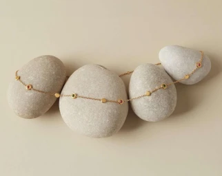 Delicate gold chain necklace with colorful gemstones draped over four smooth river stones on beige background.