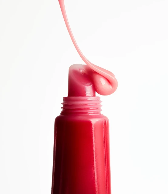 Close-up shot of a tube of lip gloss with a stream of pink liquid coming out, set against a clean white backdrop. Beauty product.