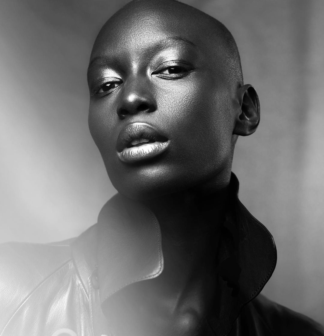 Striking black and white portrait of a bald, dark-skinned woman wearing a leather jacket, with metallic makeup and smooth skin in studio lighting.