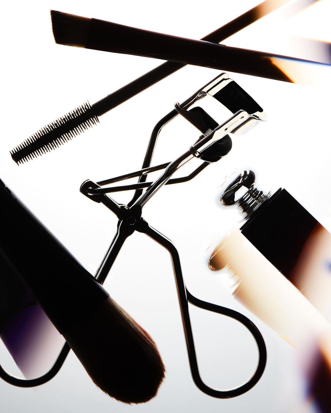 Close-up of makeup products including an eyelash curler, mascara, eyeliner, and a cosmetic brush on a bright background.
