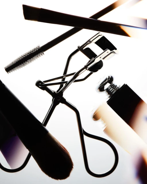 Close-up of makeup products including an eyelash curler, mascara, eyeliner, and a cosmetic brush on a bright background.