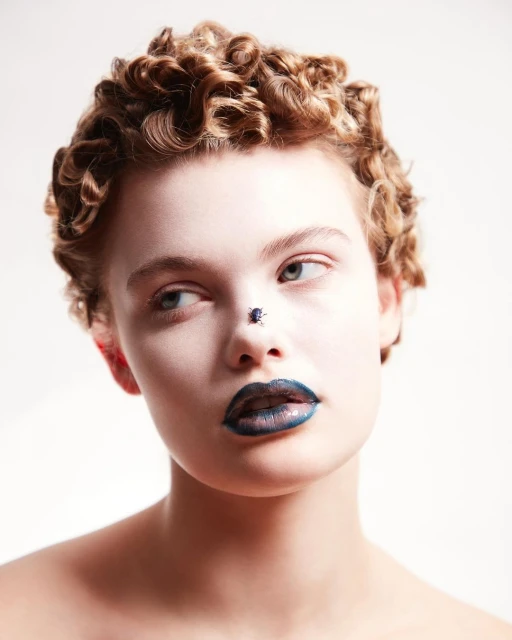 Close-up portrait of a young adult female with a blue iridescent beetle on her nose and glossy dark blue lipstick against a plain background.