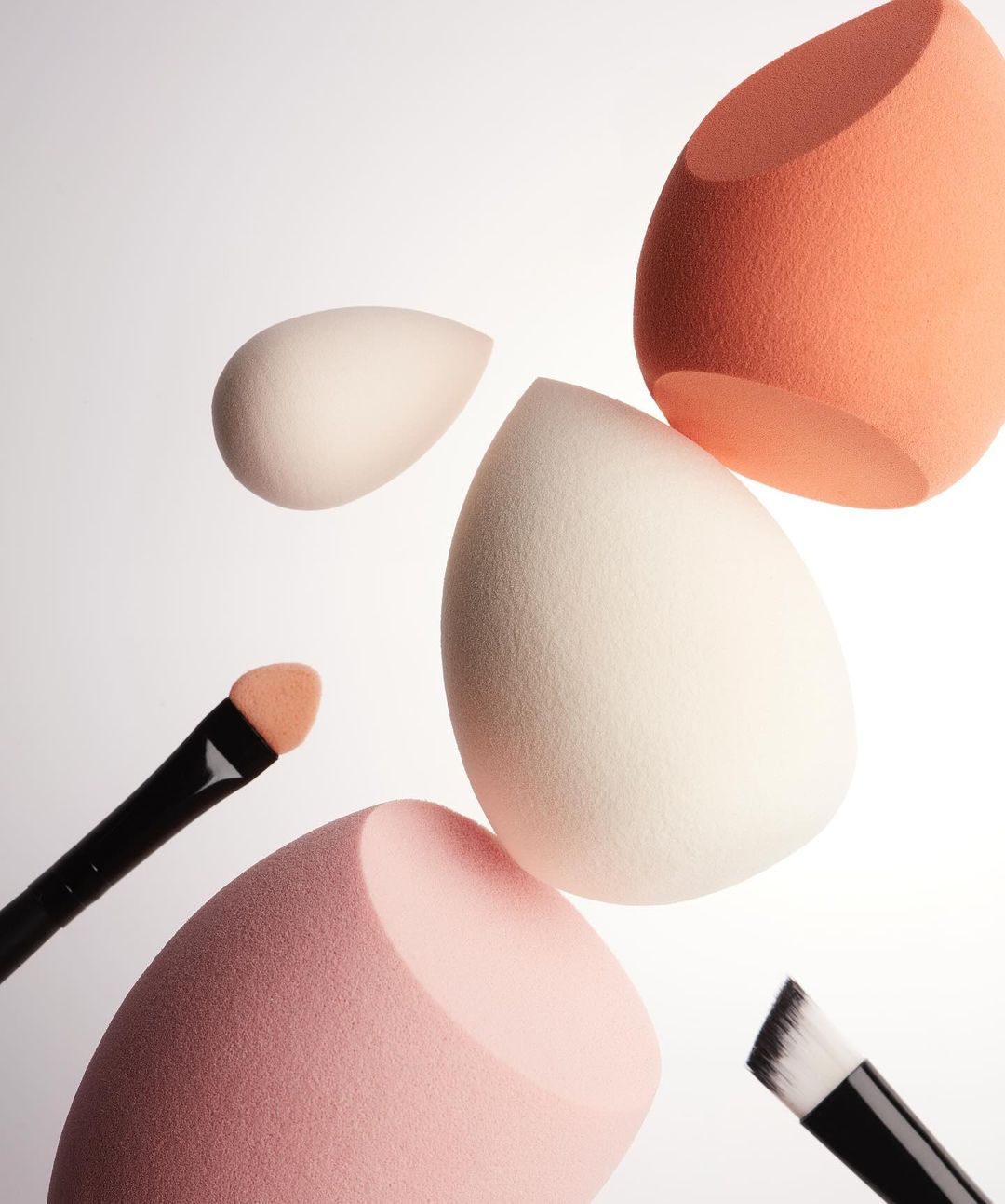 Several makeup sponges in varying colors (white, pink, orange) and sizes are arranged with makeup brushes on a white background.