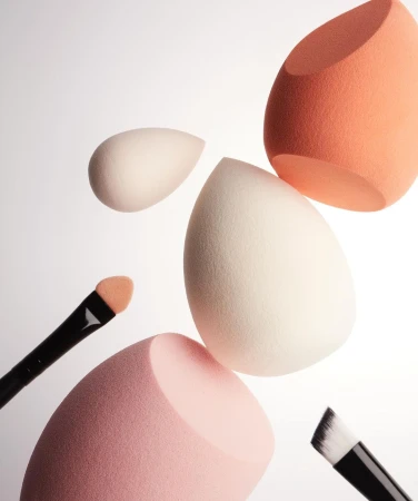 Several makeup sponges in varying colors (white, pink, orange) and sizes are arranged with makeup brushes on a white background.