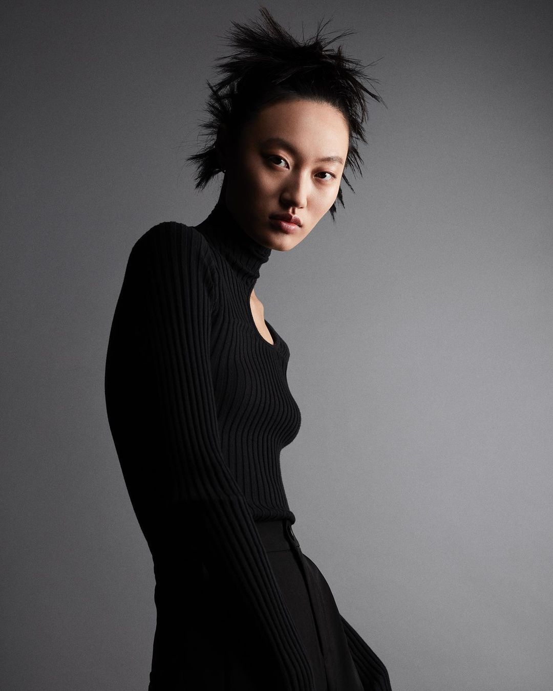 Asian female model poses in black turtleneck and slacks against a gray backdrop in a studio setting. Fashion shot, portrait orientation.