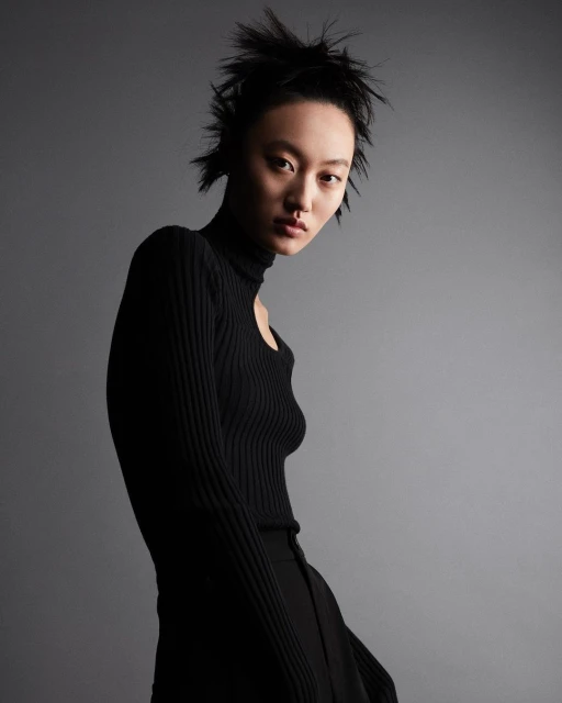 Asian female model poses in black turtleneck and slacks against a gray backdrop in a studio setting. Fashion shot, portrait orientation.
