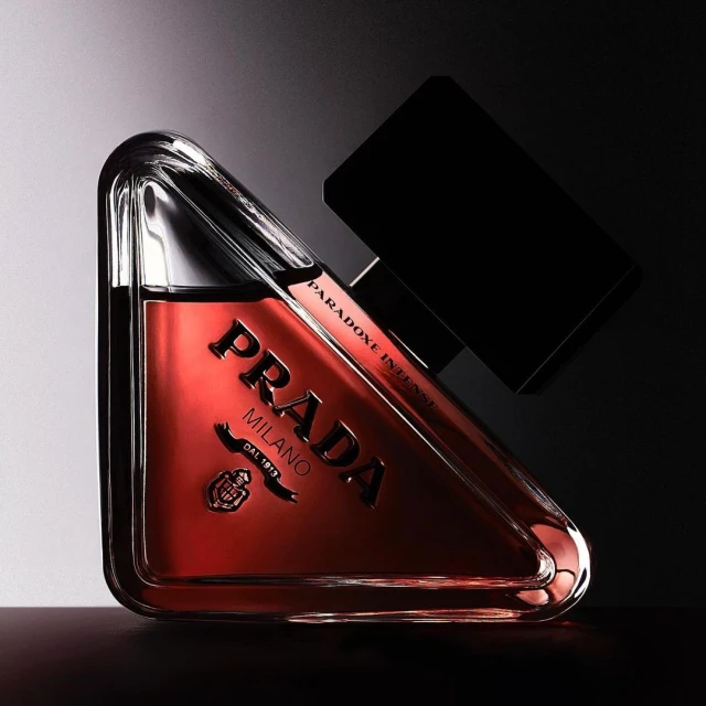 Prada Paradoxe Intense perfume bottle with a triangular shape, black lid, and dark amber liquid, displayed against a gradient backdrop.
