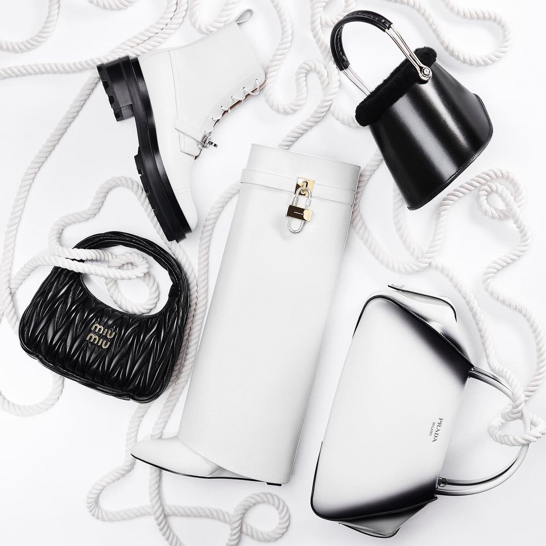 Elevated studio shot of luxury black and white designer handbags and boots arranged on a clean white background with white rope.