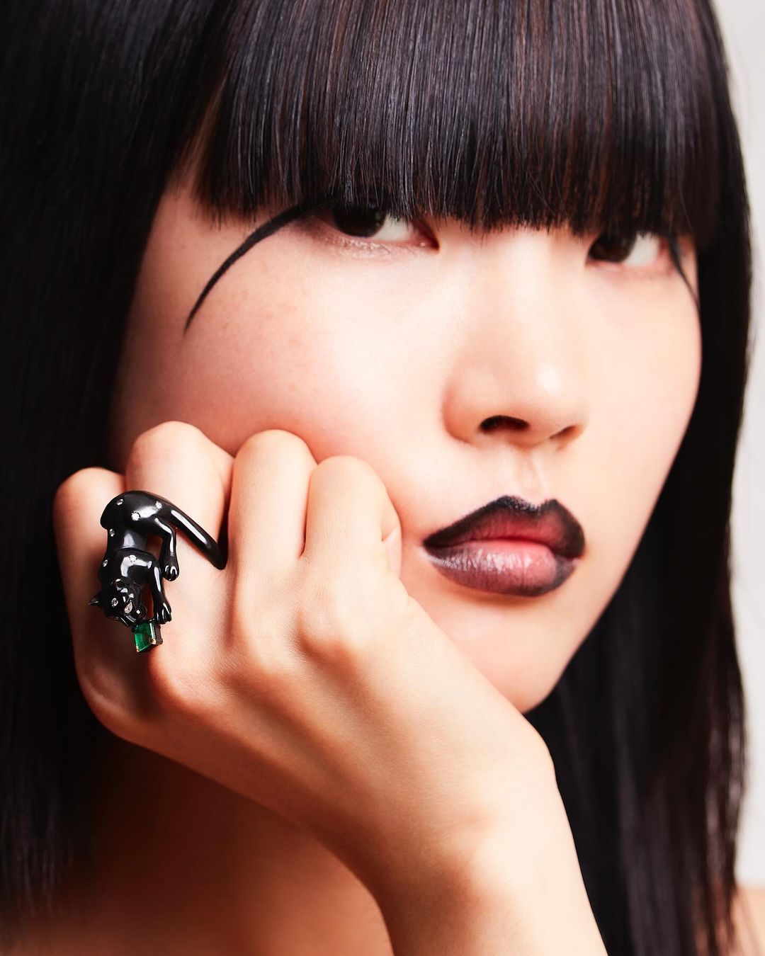 Close-up of a woman with dark hair and dramatic makeup showing a black panther ring with green gems on her finger in a beauty style shot.