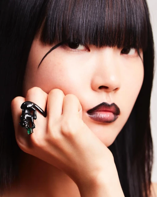 Close-up of a woman with dark hair and dramatic makeup showing a black panther ring with green gems on her finger in a beauty style shot.