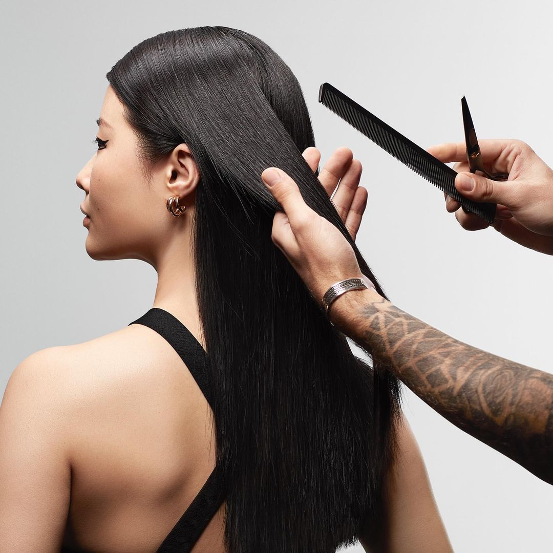 Elegant Asian woman gets her long, straight, black hair styled at a beauty salon using comb and scissors against a plain light backdrop.