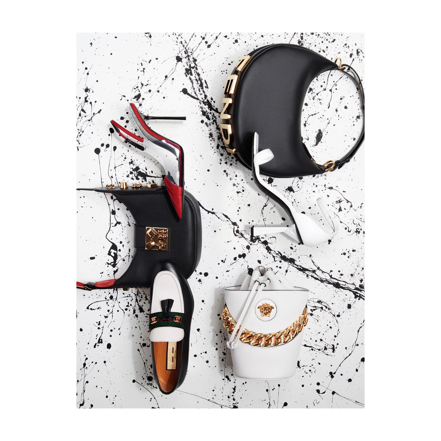 High-angle shot of luxury fashion items including handbags, high-heeled shoes, and a loafer against a white, paint-splattered background.