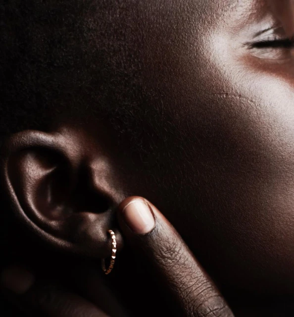 Close-up on an adult Black female face in a dark indoor setting; a gold hoop earring is visible on her ear, and her finger touches it.