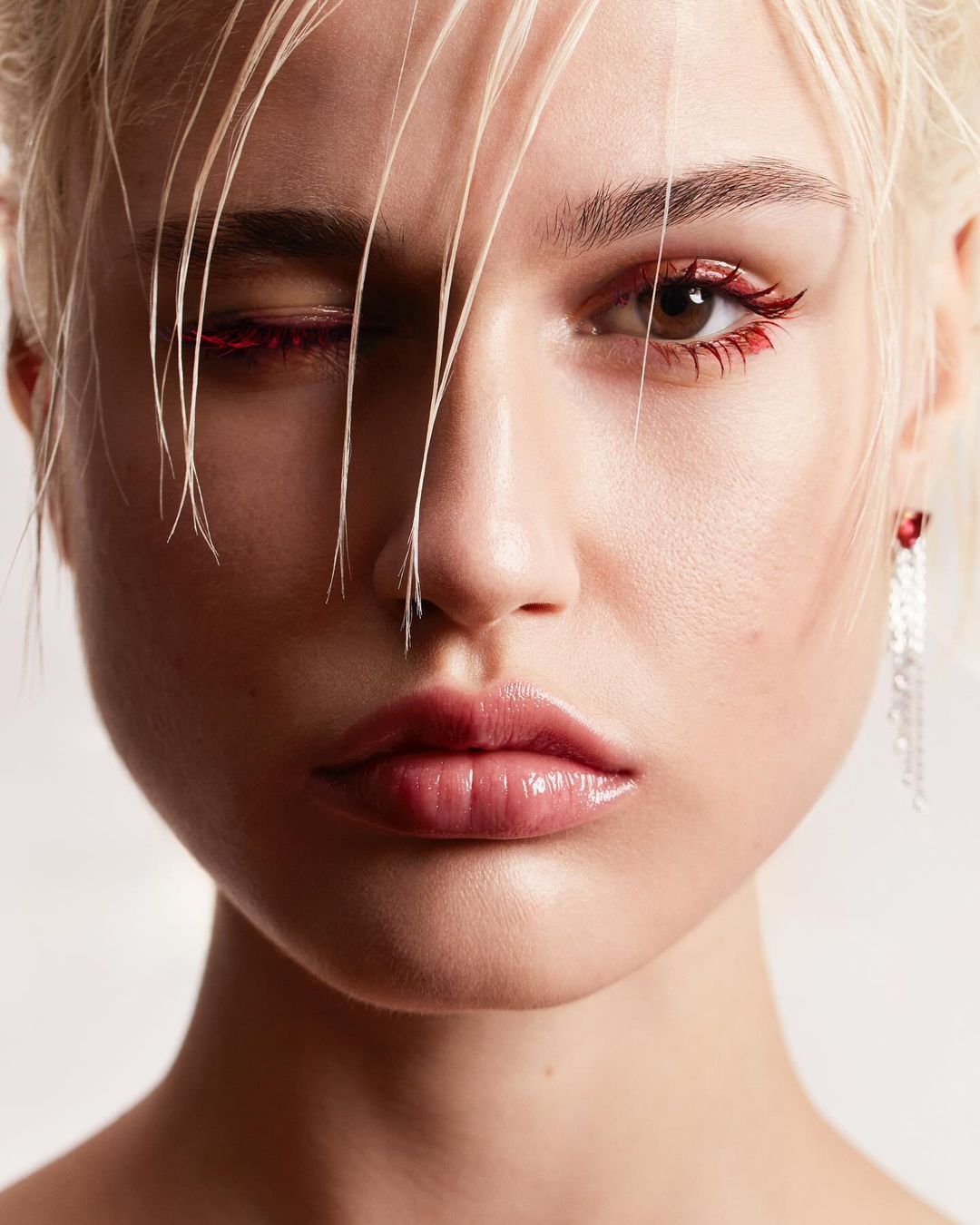 Close-up portrait of a young woman with blonde hair and red eyeliner makeup, partially obscuring one eye, set against a plain background.