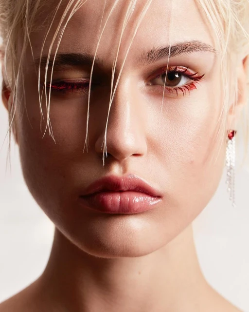 Close-up portrait of a young woman with blonde hair and red eyeliner makeup, partially obscuring one eye, set against a plain background.