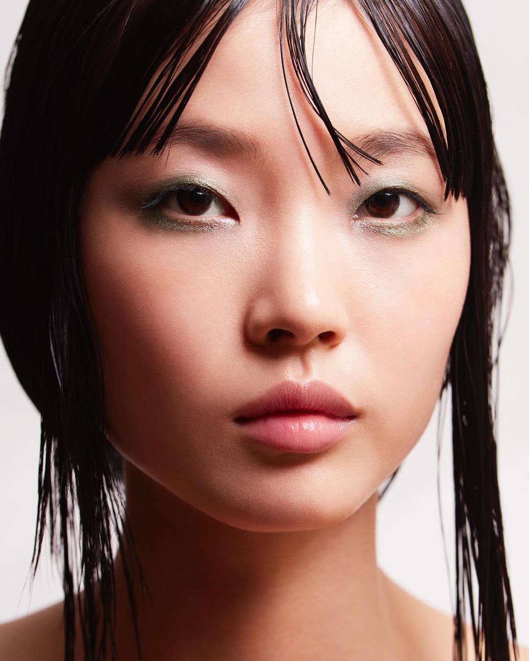 Close-up portrait of an Asian woman with sleek dark hair, minimal green eyeshadow, and glossy lips against a neutral backdrop in studio lighting.