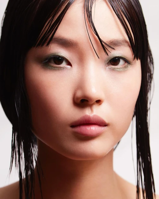 Close-up portrait of an Asian woman with sleek dark hair, minimal green eyeshadow, and glossy lips against a neutral backdrop in studio lighting.