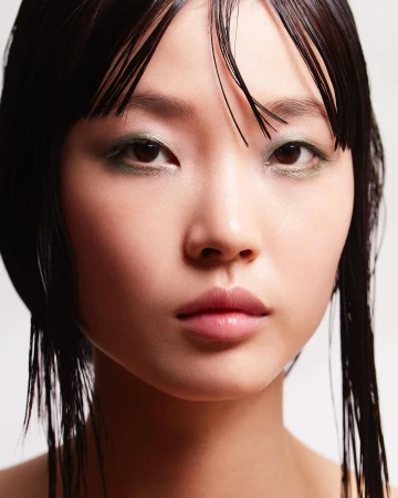 Close-up portrait of an Asian woman with sleek dark hair, minimal green eyeshadow, and glossy lips against a neutral backdrop in studio lighting.