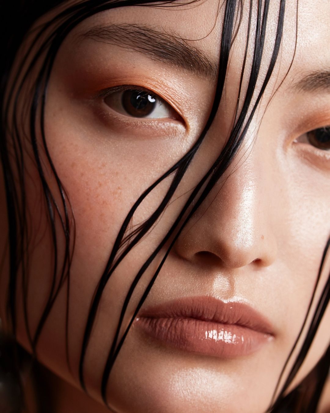 Close-up portrait of an Asian adult female with wet dark hair framing her face and glossy nude makeup, set against a dark background.