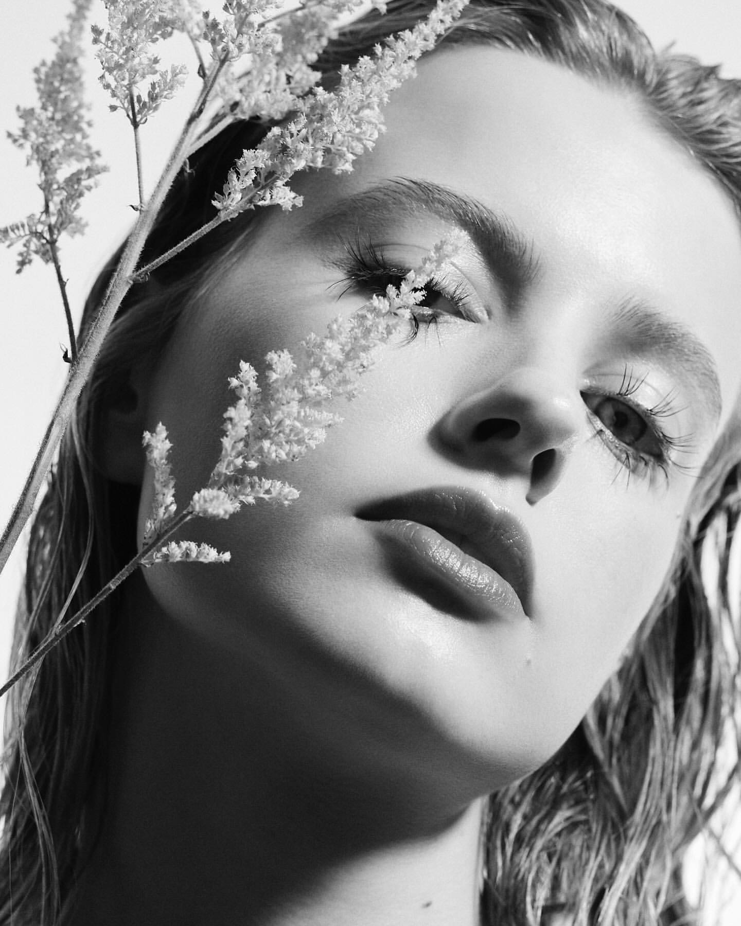 Monochrome portrait of a young woman with fair skin and light hair, styled with wet look, posing with sprigs of delicate white flowers.