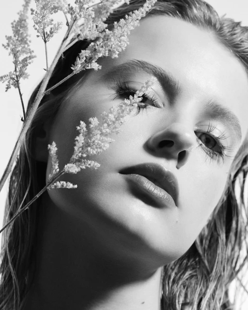 Monochrome portrait of a young woman with fair skin and light hair, styled with wet look, posing with sprigs of delicate white flowers.
