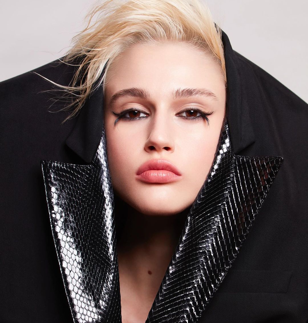 Close-up portrait of a young woman with bleached blonde hair wearing heavy black eyeliner and a snakeskin collar jacket, against a white background.