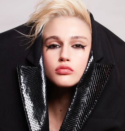 Close-up portrait of a young woman with bleached blonde hair wearing heavy black eyeliner and a snakeskin collar jacket, against a white background.
