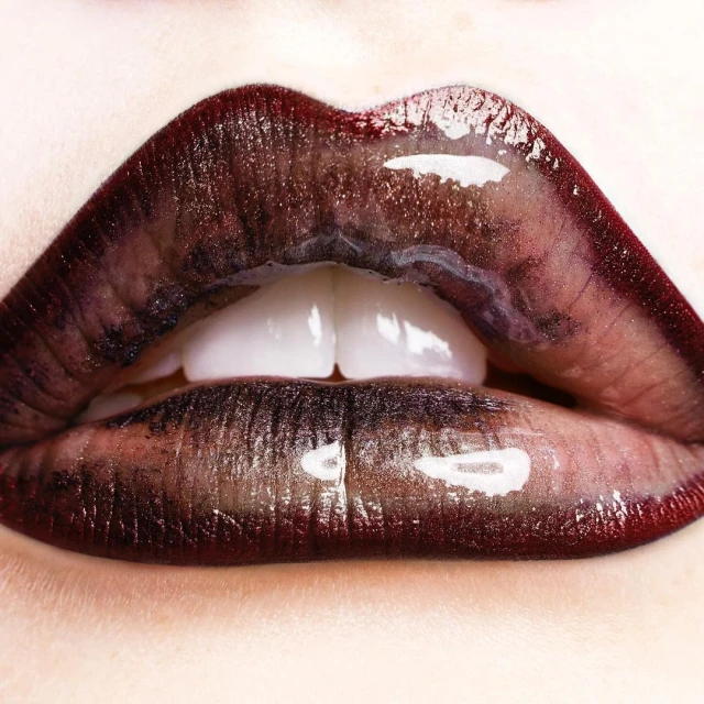 Macro close-up of human lips with glossy, dark red lipstick. The glossy texture highlights the lip's form and the teeth are partially visible.