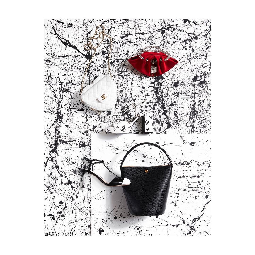 Fashion still life: Heart-shaped Chanel purse, red clutch with padlock, bucket bag, and heeled sandal against splatter-painted backdrop.