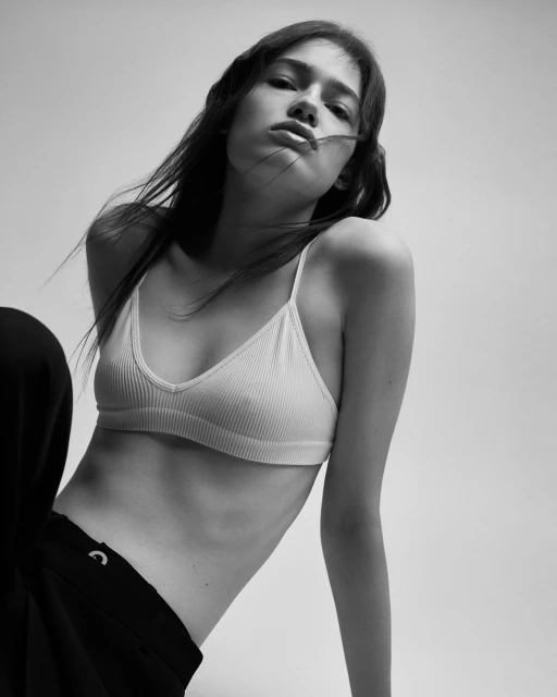 Black and white portrait of a young adult female fashion model in a white bra top and black pants against a neutral background.