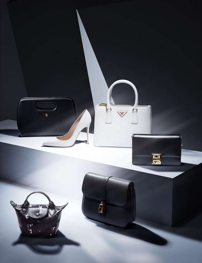 Fashion still life featuring a white stiletto heel and a collection of designer handbags in black and white, staged on geometric platforms.